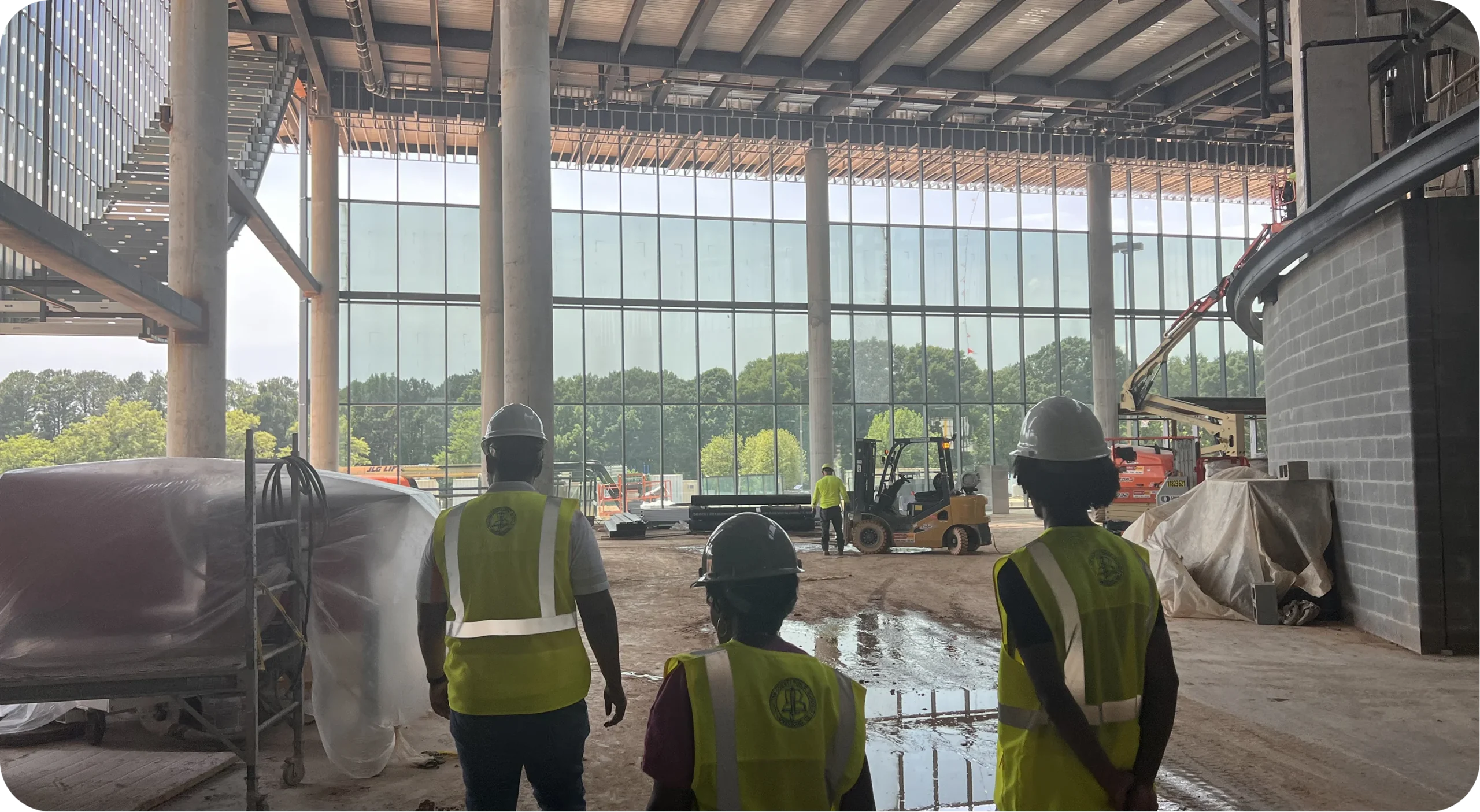 Three construction and project leaders in high-visibility safety vests and hard hats overseeing a major indoor facility development site.